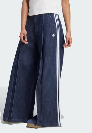 Pantalones vaqueros anchos de color azul oscuro con rayas blancas laterales y un logo de Adidas, combinados con zapatos azules y una camiseta blanca.