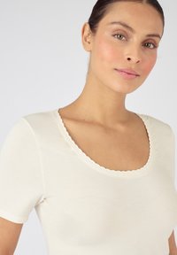 Top corto a maniche corte di un beige chiaro con scollatura a onda. Realizzato in tessuto morbido e liscio con una vestibilità aderente, offre una silhouette pulita e semplice.