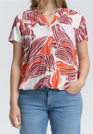 Kobieta nosząca bluzkę z krótkim rękawem w czerwone i pomarańczowe liście, w połączeniu z jasnoniebieskimi dżinsami, prawa ręka w kieszeni.