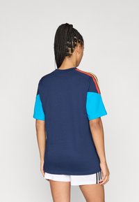 Γυναίκα με πλεξούδα, στριμμένη ώστε να κοιτάζει προς τα πίσω, φοράει ναυτικό μπλουζάκι με μπλε μανίκια και κόκκινες-κίτρινες ρίγες, συνδυασμένο με λευκές βερμούδες.