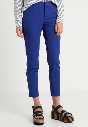 Osoba nosząca dopasowane niebieskie spodnie, czarne sandały na platformie z drewnianymi, paskowanymi podeszwami oraz białą bluzkę z długim rękawem w czarne diagonalne paski.