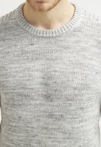 Pull gris clair tricoté avec un col rond, présentant un motif texturé et de subtiles taches plus foncées réparties sur le tissu.