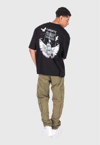Svart oversized t-skjorte med et grafisk trykk av Frihetsgudinnen med vinger, og ordet "LIBERTY" over. Olivenfargede cargo-bukser, hvite sneakers.