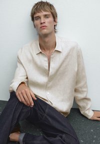 Light beige linen shirt with button front, relaxed fit, and slightly textured fabric, paired with dark denim jeans featuring light stitching.