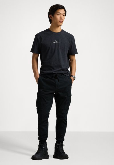 Tricou negru cu mânecă scurtă și logo alb, combinat cu pantaloni cargo negri cu buzunare laterale. Modelul poartă bocanci negri cu șiret.