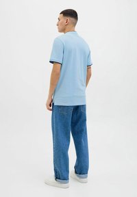 Polo shirt azzurro chiaro a maniche corte con rifiniture nere, abbinato a jeans blu a vestibilità ampia. Il modello si trova davanti a uno sfondo neutro.