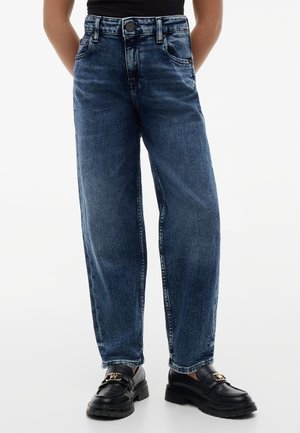 Voksen iført mørkeblå jeans med lige ben og sorte læderloafers med guldsnude, stående med hænderne bag ryggen.