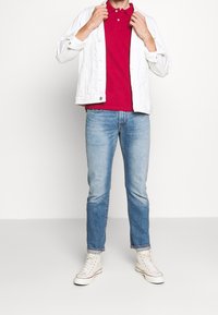 Veste blanche à boutons sur une chemise polo rouge, associée à un jean bleu clair et des baskets blanches montantes. Éléments de design simples et décontractés.