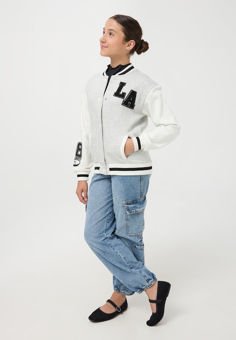 Giacca varsity grigia e bianca con "LA" in paillettes nere, polsini a costine e chiusura a bottone. Abbinata a pantaloni cargo blu chiaro e ballerine nere.