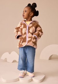 Gebreid bruin en beige jas met bloemenpatroon en roze accenten, gecombineerd met lichtblauwe wijde broekspijpen en pastelgekleurde sneakers.