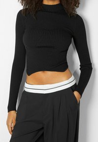 Černý žebrovaný crop top s dlouhými rukávy a vysokým límcem, zdobený trojúhelníkovým švem. Doplněno volnými černými kalhotami s bílým pasem.