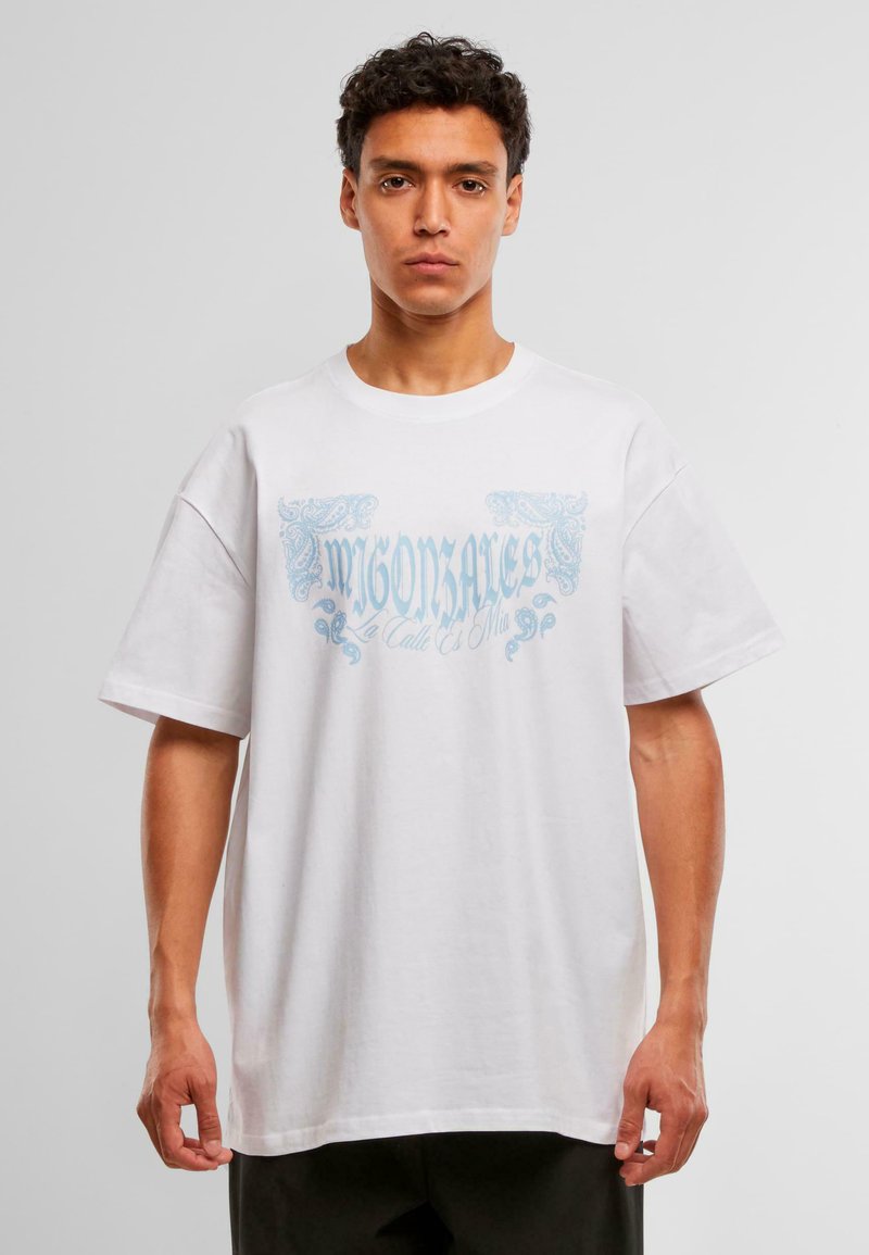 Hombre vestido con una camiseta blanca de gran tamaño con el texto y diseño decorativo en azul claro "MIGONSALES La Calle Es Mía", de pie contra un fondo sencillo.