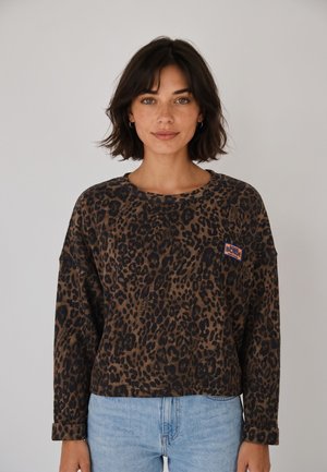 Mujer con cabello corto y oscuro que lleva una camiseta de manga larga con estampado de leopardo en marrón y negro y vaqueros azul claro, de pie frente a un fondo sencillo.