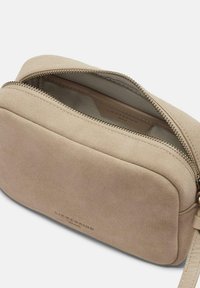 Borsa a tracolla in suede beige con chiusura a zip, forma arrotondata e design minimalista. Presenta un logo in rilievo discreto sulla parte anteriore.