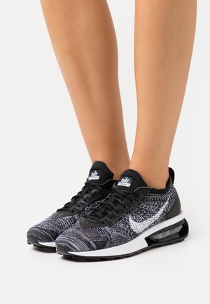 Jambes portant des chaussures de sport Nike Flyknit noir et blanc avec un amorti visible et une texture en tricot, se tenant sur une surface blanche unie.
