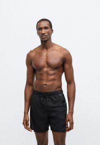 Μαύρα μαγιό σορτς με ελαστική μέση και κορδόνι, πλαϊνές διακοσμητικές ρίγες και απαλό ύφασμα. Σχεδιασμένα για άνεση και ελευθερία κινήσεων.