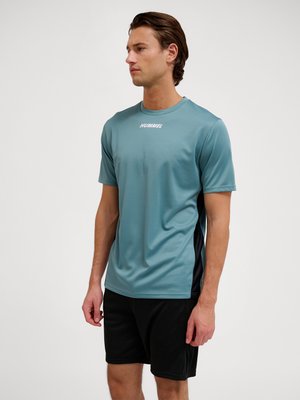 Mężczyzna noszący turkusową sportową koszulkę z krótkim rękawem z logo "HUMMEL" i czarne spodenki, stojący na jednolitym jasnym tle.