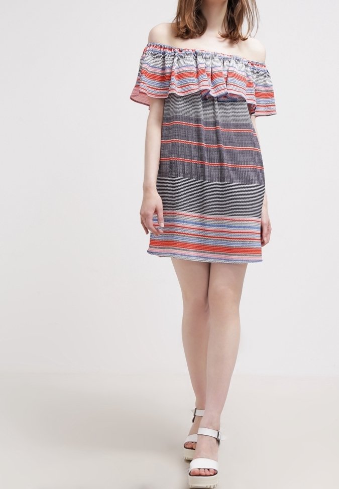 Femme portant une robe courte épaules dénudées à rayures horizontales noires, blanches, rouges et bleues, associée à des sandales blanches à plateforme.