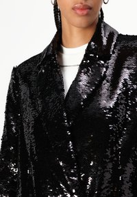 Svart paljettblazer med høyglanset overflate, med klassisk reversdesign og myk tekstur. Bæres over en hvit topp med en sølvkjede.