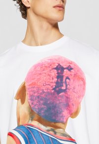Hvit bomulls t-skjorte med en fargerik illustrasjon av en person med rosa, krøllete hår på baksiden, iført en blå og rød stripete jersey.