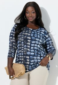 Marineblå top med abstrakte lyseblå mønstre, rund halsudskæring, trekvartærmer, kombineret med beige bukser og guldaccessories.