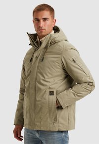 Beige vadderad jacka med huva, med dragkedja, sidofickor och en texturerad panel framtill. Inkluderar dragskoaccent.