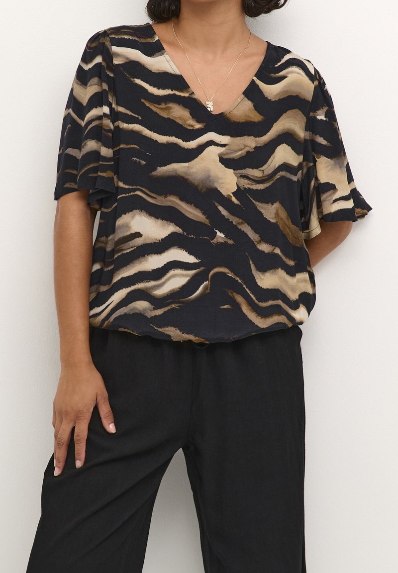 Blouse sombre avec un col en V, des manches courtes, et un motif abstrait de vagues en beige et marron, fabriquée dans un tissu lisse. Portée avec un pantalon noir.