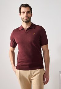 Burgundy lyhythihainen polo-paita pehmeästä kankaasta, jossa on kauluspääntie ja pieni keltainen logo vasemmassa rinnassa. Yhdistetty beigen värisiin housuihin.