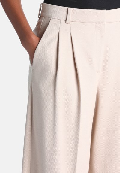 Personne à la peau foncée portant un pantalon beige plissé taille haute, main dans la poche gauche, sur un fond uni.