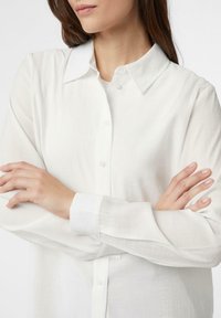 Biała koszula zapinana na guziki, wykonana z lekkiego materiału, z klasycznym kołnierzykiem, długimi rękawami i teksturowanym wykończeniem.