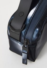 Marineblaue Tasche mit glanzfarbener Oberfläche, ausgestattet mit einem schwarzen Tragegurt, robustem Reißverschluss und Marken-Zippertab. Glatte Textur mit dezenten Nahtdetails.