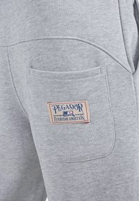Lys grå sweatpantslomme med en rød, hvid og blå Pegador mærkeetiket, der står "Everyday Ambition" syet på lommen.