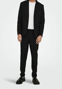 Czarny garnitur dopasowany z jednorzędową marynarką, połączony z białym t-shirtem. Posiada gładki materiał i spodnie z płaskim przodem. Czarne buty.