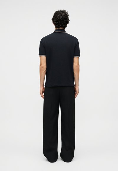 Άνδρας που στέκεται με την πλάτη γυρισμένη, φοράει μαύρο πόλο με κοντά μανίκια και λευκή επένδυση, καθώς και φαρδιά μαύρη παντελόνα, μπροστά σε απλό φόντο.