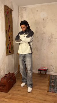 Νέος άνδρας στέκεται με σταυρωμένα χέρια φορώντας γκρι και άσπρο μπουφάν, φαρδιά τζιν και άσπρα αθλητικά παπούτσια σε δωμάτιο με ρουστίκ τοίχους.