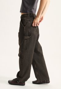 Pantalones de mezclilla marrón oscuro con un corte relajado, que cuentan con bolsillos laterales y costuras visibles. Llevados con zapatos negros y detalles de dobladillo deshilachado.