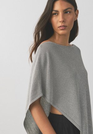 Kvinde med langt mørkt hår iført en løs, skinnende grå poncho-stil top og sorte bukser, der kigger til siden mod en ensfarvet baggrund.