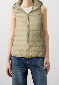 Beige Puffer-Weste mit einem Reißverschluss vorne und einer Kapuze. Verfügt über ein gestepptes Muster und hat keine Ärmel. Getragen über einem weißen Oberteil, kombiniert mit blauen Jeans.
