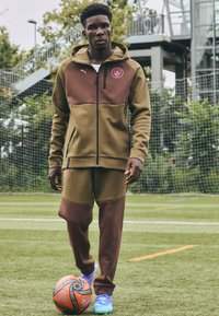 Ensemble de survêtement bicolore marron et olive, comprenant un hoodie zippé et un jogger, avec un tissu texturé et un logo brodé sur la poitrine.