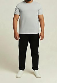 Mann trägt ein hellgraues kurzärmeliges T-Shirt, schwarze hochgekrempelte Hose und weiße Turnschuhe, steht vor einem schlichten Hintergrund.