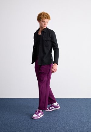 Junger Mann mit lockigem blondem Haar, der ein schwarzes Hemd, ein schwarzes Tanktop, lila Hose und lila-weiße Turnschuhe trägt und auf einem blauen Teppich steht.