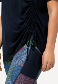 Top blu navy con dettagli laterali arricciati e cordoncino, abbinato a leggings multicolore con motivi geometrici e accenti in rete.