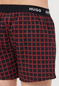 Boxers noirs avec un motif répétitif "HUGO" rouge, une ceinture élastique avec le logo "HUGO" blanc, et une texture de tissu lisse.