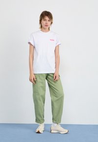 T-shirt blanche à manches courtes avec un logo rose, associée à un pantalon cargo vert. Le mannequin porte des baskets beiges, debout sur un sol bleu devant un fond clair.