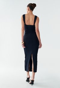 Juoda glotni midi suknelė su aptaisyta siluetu, kvadratine nugara ir šoniniu pjaustymu. Medžiaga turi subtilų blizgesį. Derinama su juodais aukštakulniais.