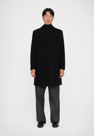 Abrigo de lana negro con diseño de un solo botón, solapas en pico, dos bolsillos frontales y corte recto, combinado con pantalones grises y zapatos negros.