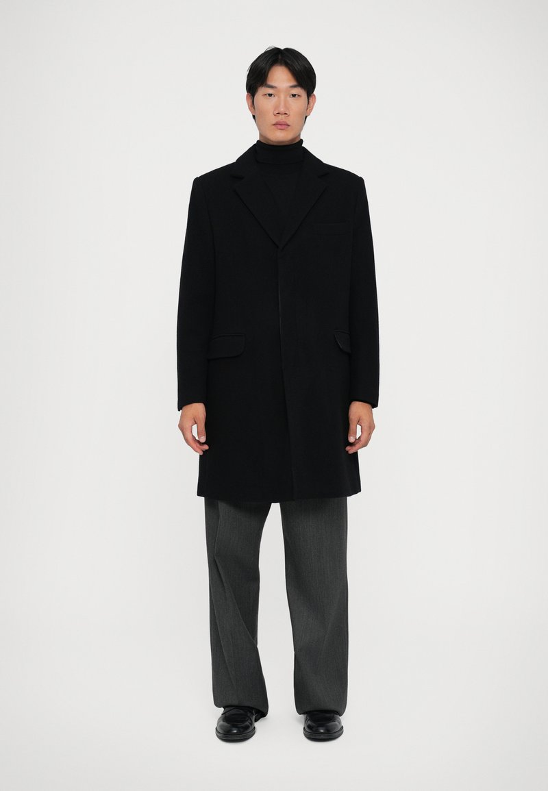 Cappotto di lana nero con design a doppio petto, revers a punta, due tasche frontali e una vestibilità dritta, abbinato a pantaloni grigi e scarpe nere.