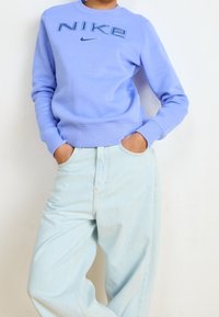 Person som bär en ljuslila Nike-tröja med logotyp, ihop med lösa ljusblå jeans, med händerna i fickorna, stående mot en vit vägg.