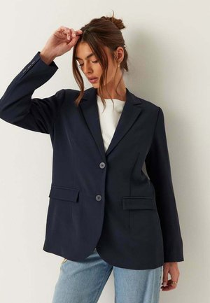 Kvinde med brunt hår samlet i en knold iført en mørkeblå blazer over en hvid top og lyseblå jeans, poserer med den ene hånd hævet nær panden.