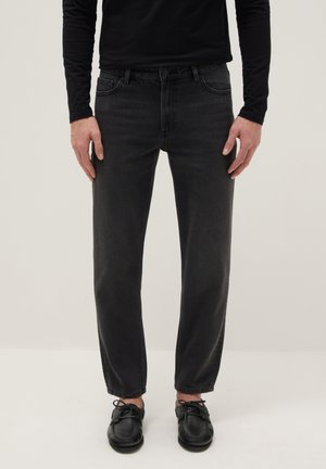 Personne portant un jean noir slim, une chemise noire à manches longues et des chaussures noires à lacets, debout devant un fond clair uni.